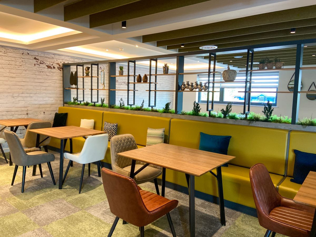 A modern cafe interior featuring wooden tables and a variety of seating options, including upholstered chairs. Greenery accents the space, with shelves displaying decorative items in the background. Soft lighting enhances the inviting atmosphere.